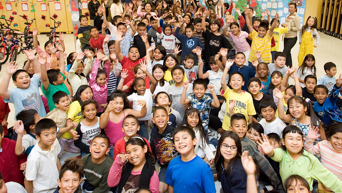A classroom of elementary school children cheering at the camera, with bicycles and adults behind them